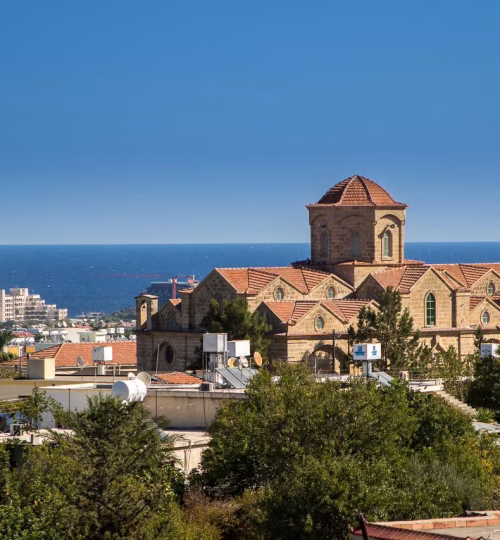 A scenic view of a historic stone church with red-tiled roofs, surrounded by greenery. Overlooks the Mediterranean Sea with modern city buildings in the distance under a clear blue sky.