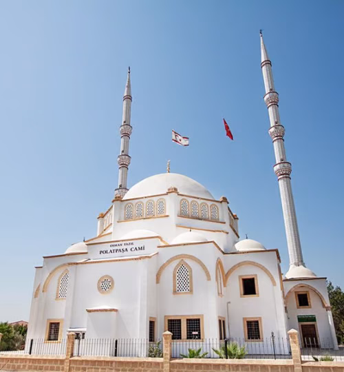 The Polatpaşa Mosque, a stunning white structure with golden accents, featuring two tall minarets and a central dome. Set under a clear blue sky with flags flying above.