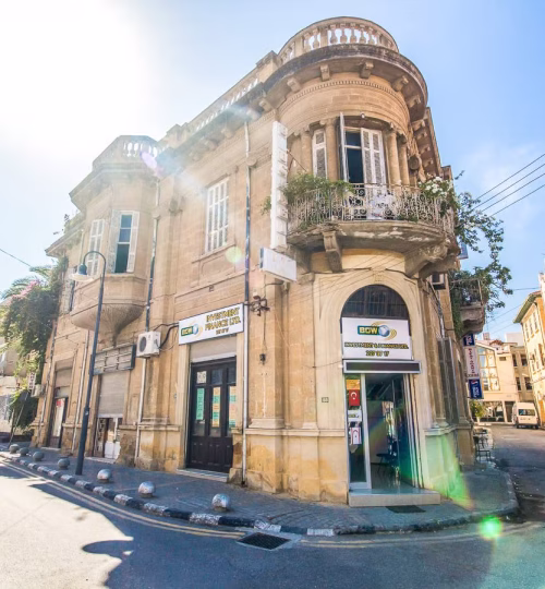 A charming historic building with ornate balconies and arched windows, located on a sunny street corner. The ground floor houses a local business with a classic architectural style.