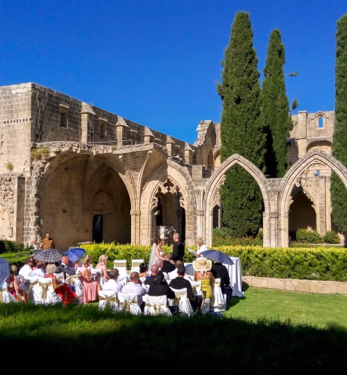 A scenic outdoor wedding ceremony set against the backdrop of a historic stone abbey. Guests are seated on a lush green lawn, surrounded by Gothic arches and tall cypress trees under a bright blue sky.